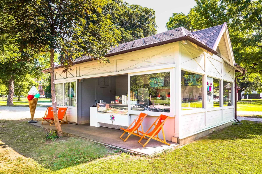 Der Eissalon im Augarten in Graz: weißer Pavillon mit orangen Liegestühlen im Park