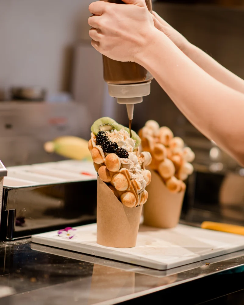 Bubble waffle with ice cream and fruit being topped with sauce