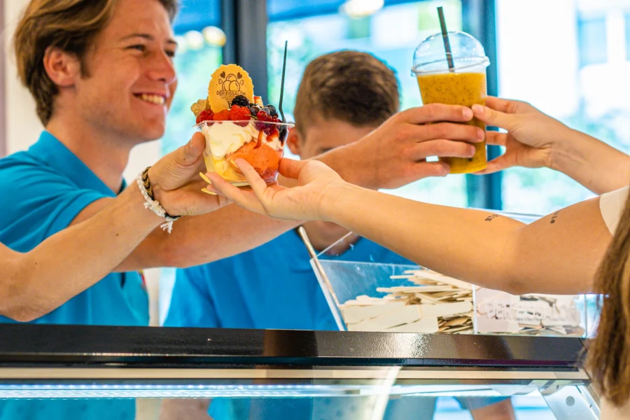 Lächelnder Mitarbeiter reicht bei Der Eissalon in Graz einen Eisbecher und einen Smoothie über die Theke